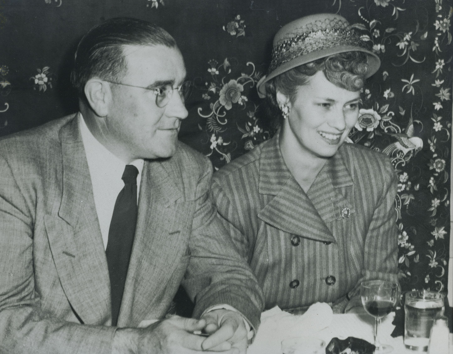 Black and white image of man in suit and woman in hat and jacket sitting at table